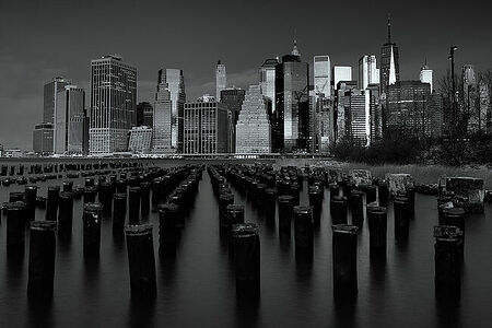 This black and white photograph captures the serene beauty of an urban skyline, juxtaposed against the historic wooden pilings in the foreground. It offers a moment of tranquility amidst the bustling city life and reflects a blend of modern architecture with historical remnants, ideal for contemporary decor themes.