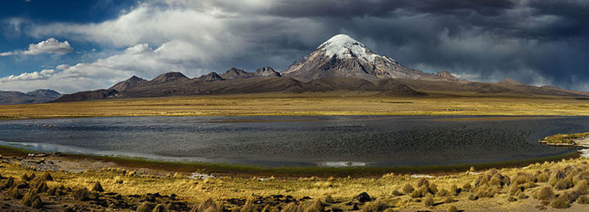 This powerful image captures the looming presence of Mount Sajama, Bolivia’s highest peak, under the dramatic dance of storm clouds in the sky. The tranquil waters below reflect the mountain’s snowy cap, contrasting with the arid landscape and the flock of flamingos gracing the wetlands. The scene is a dynamic display of nature’s contrasts, from the arid ground to the lush mountain, crowned by the tumultuous sky.
