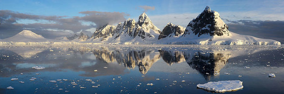 As the sun dips towards the horizon, its golden light bathes the Antarctic peaks, captured here in a moment of stunning clarity. The mirror-like waters offer a perfect reflection, doubling the majesty of the snow-capped mountains and the scattered ice that adorns the sea. This panoramic shot is a breathtaking showcase of the polar landscape, where the harmony of nature’s grandeur and tranquility is eternally preserved.