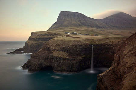 This serene image features the iconic Gasadalur waterfall on the Faroe Islands, flowing gracefully into the ocean. Above, the quaint village of Gasadalur nestles against the dramatic backdrop of towering mountains, shrouded in a soft haze, encapsulating the tranquil and isolated beauty of this unique location.