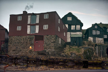 This image beautifully captures the reflection of traditional Faroese houses with grass roofs on the water’s surface. The scene, with its warm light from a window and smoke rising from a chimney, evokes a cozy, lived-in atmosphere amidst the historic architecture of the Faroe Islands.