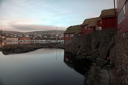 This image depicts a serene evening at a harbor in the Faroe Islands, with traditional red houses boasting grass roofs reflecting in the calm waters. The distant view of the town provides a vibrant backdrop, while a lone figure enjoying the view embodies the peaceful solitude that the islands offer.