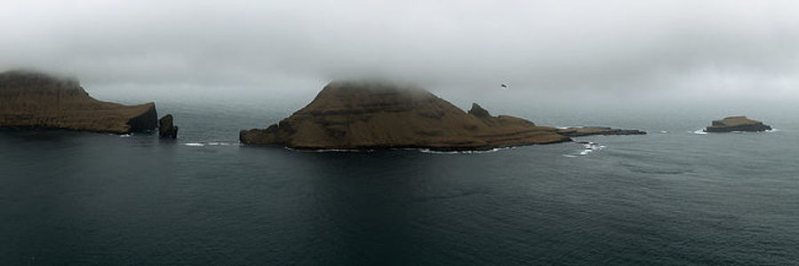 A panoramic view of the Faroe Islands, where the majestic cliffs and peaks are shrouded in mist, evoking a sense of mystery and untouched wilderness. The North Atlantic Ocean surrounds the archipelago, its waters gently lapping against the rugged shorelines. A solitary bird in flight accentuates the expansive solitude of this remote paradise.