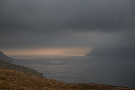 This captivating image showcases a serene view of the Faroe Islands, with the radiant light peeping through the heavy clouds, casting a warm glow over the North Atlantic Ocean. The rugged landscape foreground contrasts with the peaceful sea, creating a perfect harmony of nature’s drama and tranquility.