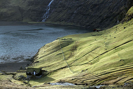 Живописная сцена разворачивается с традиционным домом на Фарерских островах, уютно расположенным в зеленой долине. Фоном для него служит падающий водопад, усиливающий ощущение уединения и гармонии с природой. Это изображение захватывает суть фарерских пейзажей, идеально подходящих для тех, кто ищет визуальное погружение в нетронутую природу.