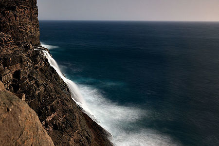 An extraordinary view of a waterfall plunging into the ocean from the steep cliffs of the Faroe Islands. The sheer force of the water creates a misty veil against the deep blue of the North Atlantic, illustrating the raw power and untouched beauty of the islands’ landscapes.