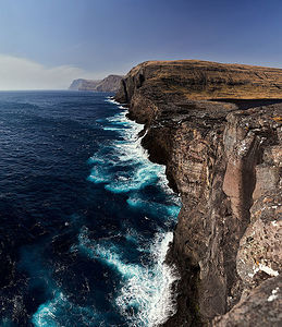 This striking image captures the raw beauty of the Faroe Islands’ cliffside, where the azure waves of the North Atlantic crash against the ancient, rugged coastline. The interplay of light and shadow across the cliffs accentuates the wild and untamed essence of this breathtaking landscape.