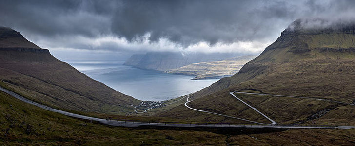 This panoramic image captures the breathtaking view of winding roads carving through the dramatic landscapes of the Faroe Islands. Clouds hug the towering cliffs which stand guard over the tranquil village and the vast sea, conveying a sense of remote beauty and the adventurous spirit of the islands.