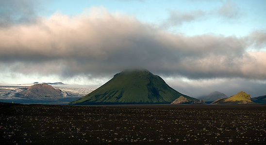 This serene landscape captures the essence of the Fjallabak Nature Reserve in Iceland, where green mountains rise majestically above the volcanic plains. The central peak, shrouded in a cloak of clouds, is illuminated by a break in the overcast sky, casting a spotlight on its lush slopes. This contrast of dark volcanic earth and vibrant greenery under the Nordic sky epitomizes the untouched wilderness of Iceland.