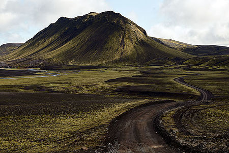 The open road beckons in this captivating image, where a meandering dirt path cuts through the heart of Iceland’s volcanic terrain, leading towards a majestic green-clad mountain. The interplay of light and shadow cast by the low-hanging sun accentuates the undulating hills and textured ground, inviting the viewer to embark on a journey through this raw and enchanting landscape.