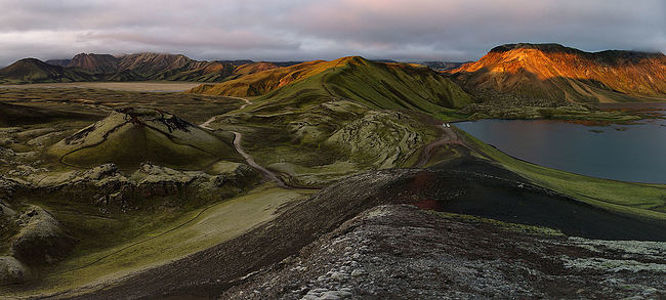 As dawn’s first light sets Landmannalaugar’s rhyolite hills ablaze, the landscape transforms into a tapestry of vibrant hues. This panoramic view captures the surreal beauty of the area, with its winding paths leading to a tranquil lake, cradled by mountains that stand as centuries-old sentinels. The contrasting textures and colors of the mossy hills, volcanic ash, and glowing peaks encapsulate the fiery heart of Iceland’s highlands.