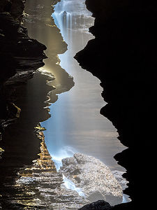 Captured in a serene cave near Pokhara, this photograph showcases the ethereal beauty of a hidden waterfall, as sunlight filters through the crevices, illuminating the cascading waters and rocky surfaces in a dance of light and shadow.