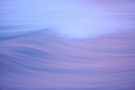 This image offers a mesmerizing view of a solitary wave captured with a long exposure technique, which smoothes its motion into a hazy blend of purple and blue hues, evoking a sense of calm as day gives way to night.