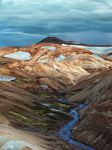 Откройте для себя потрясающий пейзаж Керлингарфьёлля - геотермального чуда на Исландском нагорье. На этом снимке запечатлен захватывающий дух контраст между риолитовыми горами, их яркими цветами от насыщенного красного до золотисто-желтого, и сверкающими пятнами снега, покоящимися в расщелинах. Меандрирующая река придает снимку безмятежный голубой оттенок, создавая гармоничное сочетание цветов и текстур.