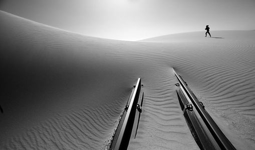 На черно-белой фотографии изображена одинокая фигура, бредущая по просторам покрытых рябью песчаных дюн, за которыми виднеются заброшенные рельсы, уходящие вдаль. Снимок вызывает чувство одиночества и масштабности пустыни, подчеркивая ее суровую красоту и созерцательную тишину.
