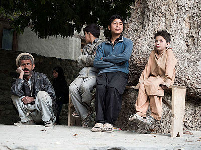 In the stillness of a Pakistani village, life pauses under the venerable branches of the village tree. Here, a solitary elder dozes in a moment of repose, his weathered features softened by slumber. Nearby, the quiet is gently pierced by the hushed interplay of youth; a boy, seated on a makeshift bench, steals a curious glance at a girl passing by, her presence weaving a silent narrative of life’s simple ebb and flow. This photograph is a serene observation of the intergenerational tapestry and the enduring bonds of a close-knit community.