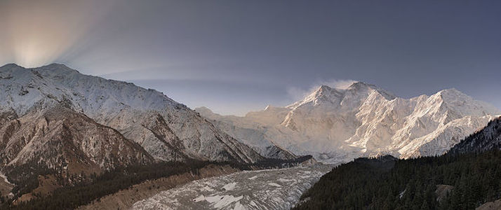 The break of dawn unveils the silent grandeur of Nanga Parbat, with the first sun rays spilling over its snowy mantles. This panorama captures the mountain in its tranquil awakening, the golden light accentuating the textures of its formidable slopes. Witness the serene moment as daybreak breathes life into the ’Killer Mountain’, a spectacle of nature’s splendor.