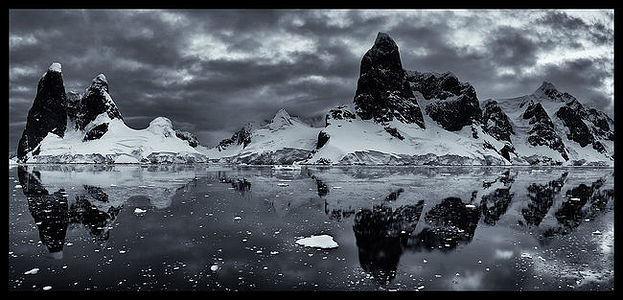 This black and white photograph captures the dramatic essence of the polar landscape, with rugged peaks rising like monoliths against a textured sky. The reflections on the glassy surface of the water add a dimension of symmetry, enhancing the stark contrasts and the timeless allure of the Antarctic’s wild beauty. The image evokes a sense of solitude and the enduring power of nature in its most elemental form.