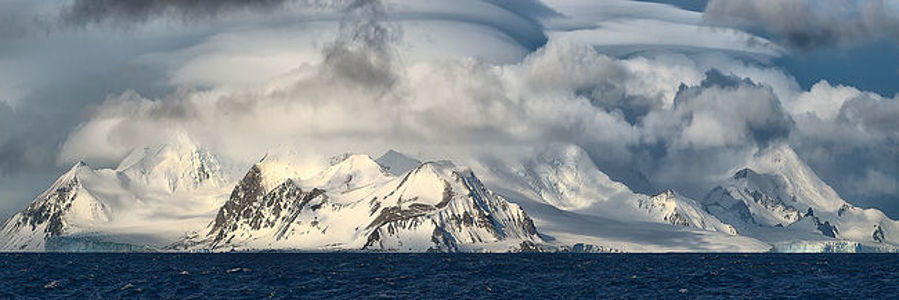 Танец света и тени на вершинах Антарктиды, где суровые горы встречаются с бурным небом. Темные клубящиеся облака, созданные жестокими полярными ветрами, подчеркивают величие покрытых снегом вершин. На этом панорамном снимке запечатлен контраст между бушующим синим морем и безмятежной белизной пейзажа - свидетельство необработанности и благоговейной мощи самой южной дикой природы Земли.