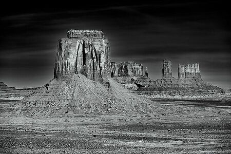 A dramatic black and white rendering of Monument Valley, capturing the raw and rugged beauty of the towering sandstone monoliths. The play of light and shadow brings out the textures and layers of the landscape, emphasising the solitude and majesty of the area.