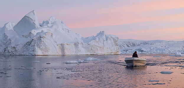 The photograph depicts a serene moment in Greenland’s arctic waters, as a boat navigates through the calm sea amidst towering icebergs under a soft twilight sky.