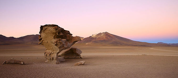The Altiplano of Bolivia presents a breathtaking tableau at dusk, where the iconic eroded rock stands as a natural sculpture against the backdrop of the majestic Andean mountains. The last rays of the sun cast a warm glow over the landscape, highlighting the unique geology of the region. The silence of the desert amplifies the solitary beauty of the rock formation, a sentinel in the vast emptiness.