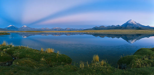 As twilight descends over the Andes, the calm waters of this high-altitude lake become a mirror, reflecting the majesty of the snow-capped peaks. The grassy banks offer a soft contrast to the rugged mountains, while the blue hues of dusk cast a tranquil ambiance over the scene. This moment of serene beauty highlights the pristine ecosystem and the breathtaking landscapes that are characteristic of the Andean region.
