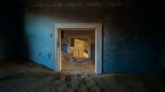 A haunting image of an abandoned house in Kolmanskop, Namibia, where the desert sands are slowly reclaiming the once-thriving diamond mining town. The decaying blue walls and the cascading dunes create a striking juxtaposition between human history and the relentless forces of nature.