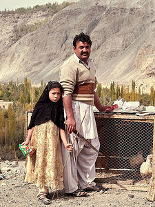 his poignant image captures a moment in the daily lives of a father and daughter in Pakistan’s mountainous region. The father, dressed in traditional attire, stands beside his daughter, her expression a mix of curiosity and wariness. Behind them, the rugged landscape of Pakistan serves as a testament to the resilient spirit of its people. The photograph conveys a sense of continuity and the enduring bonds of family against the backdrop of their ancestral land.