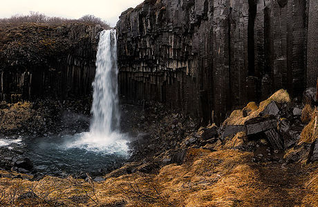 Captured in the Skaftafell National Park, this stunning photograph of Svartifoss, known for its distinctive basalt column formations, epitomizes the raw beauty of Icelandic nature. The waterfall’s icy cascade contrasts with the dark hexagonal columns and the golden brush around, creating a mesmerizing spectacle. The dark, moody sky adds a dramatic backdrop to the falls, enhancing its majestic presence.