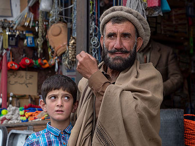 An authentic capture of a bearded elder in traditional attire with a young boy beside him, set against a backdrop of a local market, showcasing a moment of generational bonding and cultural richness.