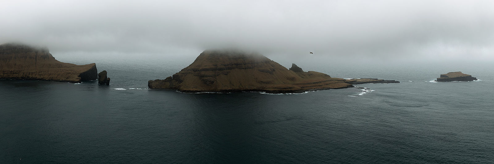 Панорамный вид на Фарерские острова, где величественные скалы и вершины скрыты в тумане, вызывая чувство таинственности и нетронутой дикой природы. Воды Северной Атлантики омывают архипелаг, нежно обтекая его изрезанные берега. Одинокая птица в полете подчеркивает безграничную уединенность этого отдаленного рая.