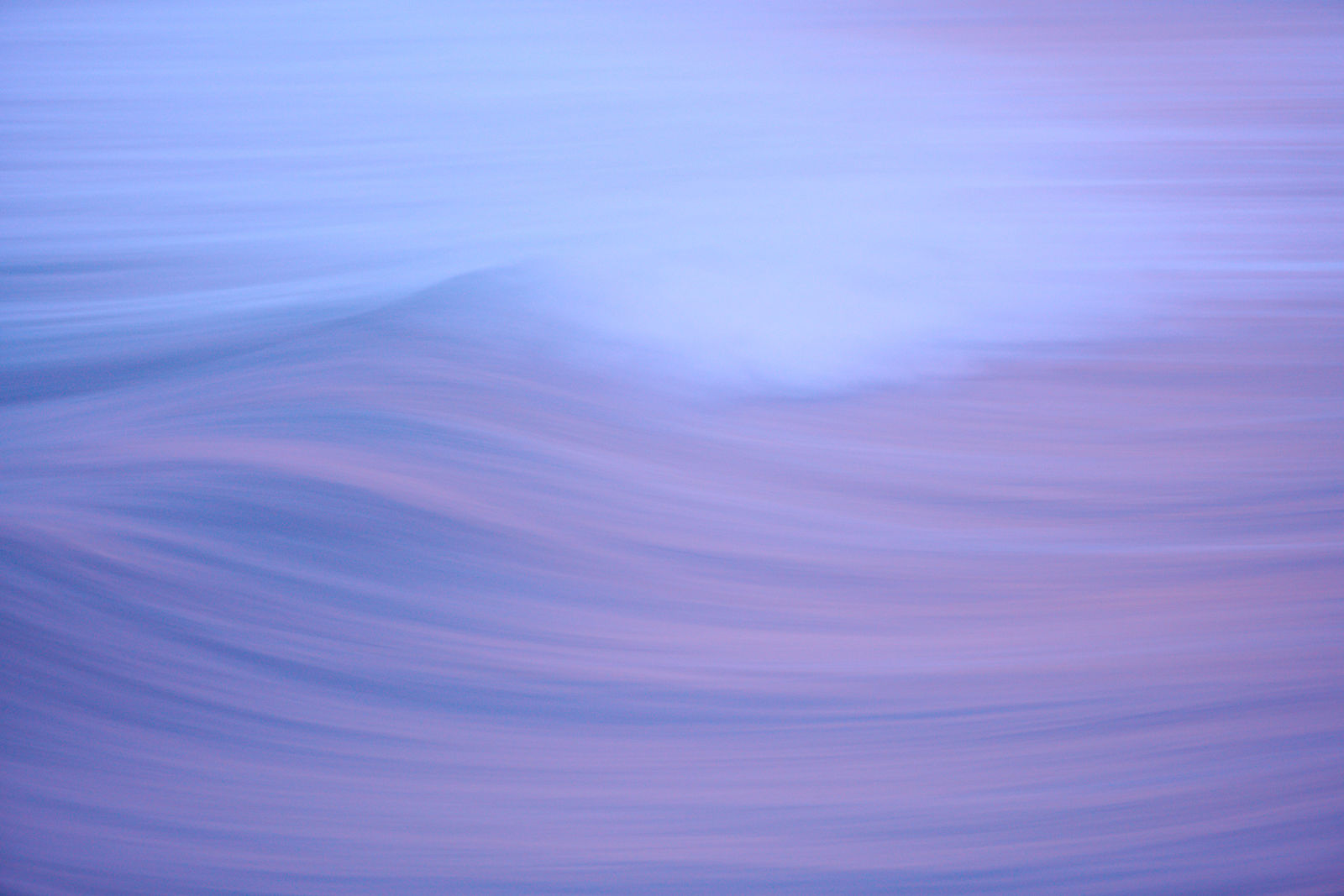 This image offers a mesmerizing view of a solitary wave captured with a long exposure technique, which smoothes its motion into a hazy blend of purple and blue hues, evoking a sense of calm as day gives way to night.